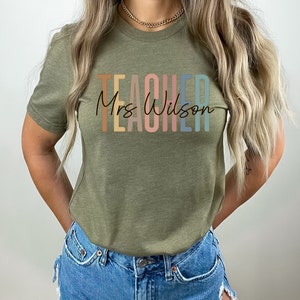 K&ouml;nnte beinhalten: Ein gr&uuml;nes T-Shirt mit dem Text "TEACHER" in Regenbogenfarben und "Mrs. Wilson" in schwarzer Schrift darunter.