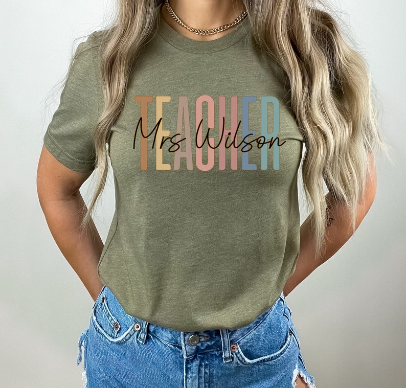 K&ouml;nnte beinhalten: Ein gr&uuml;nes T-Shirt mit dem Text "TEACHER" in Regenbogenfarben und "Mrs. Wilson" in schwarzer Schrift darunter.