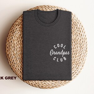 May include: Dark grey t-shirt with the text "Cool Grandpas Club" in white script. The shirt is folded and placed on a woven, circular placemat. The words "Dark Grey" are printed in a bold font at the bottom left.