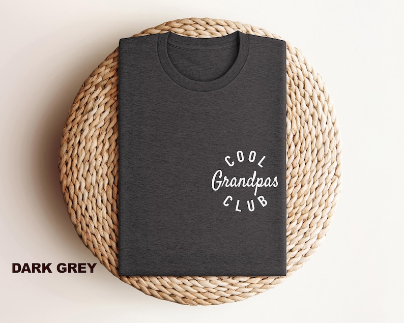 May include: Dark grey t-shirt with the text "Cool Grandpas Club" in white script. The shirt is folded and placed on a woven, circular placemat. The words "Dark Grey" are printed in a bold font at the bottom left.