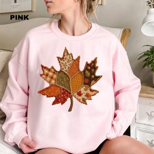 Pode incluir: Uma camisola de gola redonda rosa claro com um grande gr&aacute;fico central de uma folha de &aacute;cer. A folha &eacute; composta por v&aacute;rios padr&otilde;es de tecido estilo patchwork em tons de laranja, castanho, verde e creme, com costuras vis&iacute;veis nas bordas.