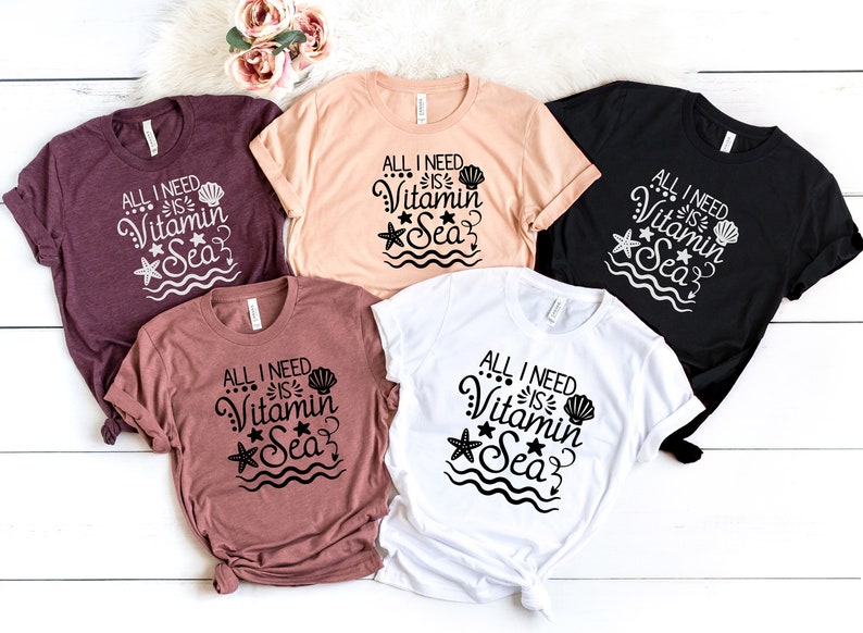 May include: Five t-shirts in different colours with the text "All I need is Vitamin Sea" in black lettering with a seashell, starfish, and wavy lines.