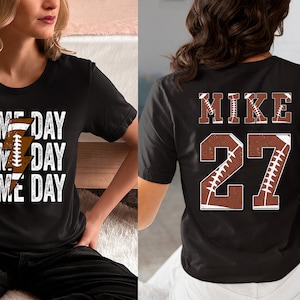 May include: Two black t-shirts with a football theme. One shirt has the text "GAME DAY" repeated three times with a lightning bolt and football graphic. The other shirt has the text "MIKE 27" with a football graphic.