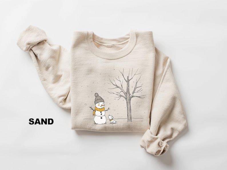 May include: A beige sweatshirt with a snowman, a small dog, and a bare tree printed on the front. The text "SAND" is printed on the bottom left corner of the sweatshirt.