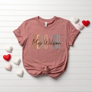 K&ouml;nnte beinhalten: Ein pinkfarbenes T-Shirt mit dem Wort "TEACHER" in Regenbogenfarben und dem Text "Mrs. Wilson" in schwarzer Schrift.