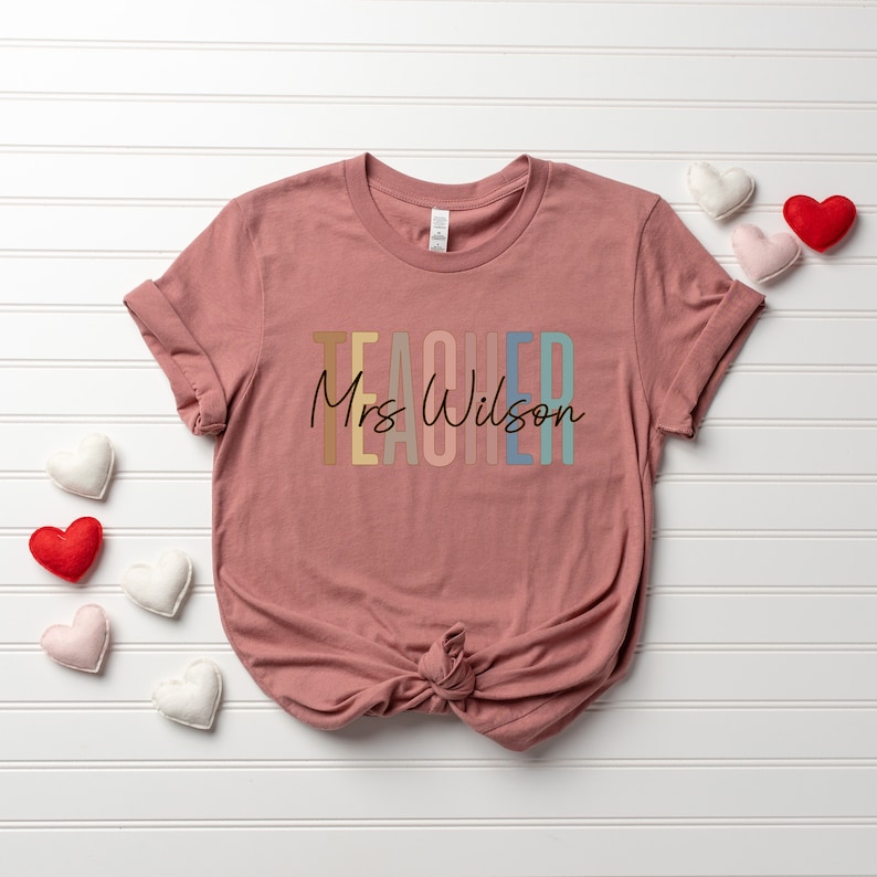 K&ouml;nnte beinhalten: Ein pinkfarbenes T-Shirt mit dem Wort "TEACHER" in Regenbogenfarben und dem Text "Mrs. Wilson" in schwarzer Schrift.