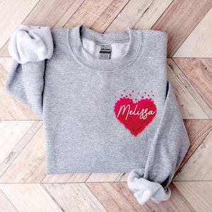 May include: A gray sweatshirt with a red heart made of smaller hearts. The heart has the name "Melissa" written inside it.