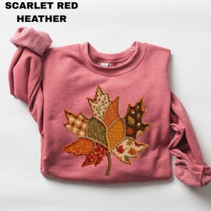 Pode incluir: Uma camisola de cor vermelho escarlate mesclado com uma grande aplica&ccedil;&atilde;o de folha de &aacute;cer ao centro. A folha &eacute; constru&iacute;da a partir de v&aacute;rios retalhos de tecido em tons de laranja, castanho, verde e creme, com padr&otilde;es de xadrez e pontos.