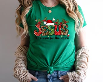 Puede incluir: Camiseta verde con un diseño navideño que presenta la palabra "JESUS" en letras coloridas con un gorro de Papá Noel y el texto "La razón de la temporada".