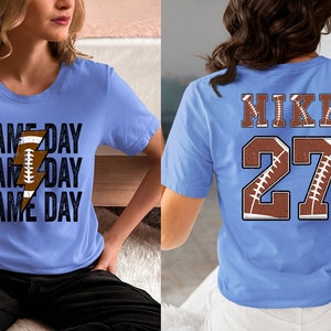 May include: Two light blue t-shirts. One shirt has the text "GAME DAY" repeated three times with a lightning bolt and football graphic. The other shirt has the text "MIKE" and the number "27" with a football graphic.