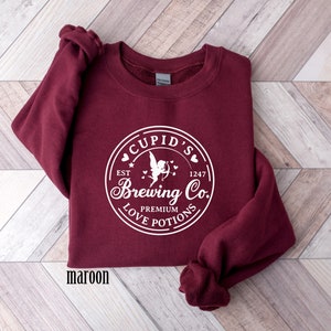 May include: Maroon sweatshirt with a white graphic of a circle with the text "Cupid's Brewing Co." and a cupid with an arrow. The text "Est 1247" is above the cupid and the text "Premium Love Potions" is below the cupid.