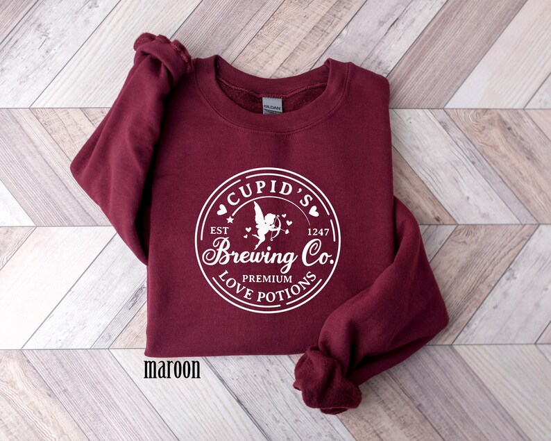 May include: Maroon sweatshirt with a white graphic of a circle with the text "Cupid's Brewing Co." and a cupid with an arrow. The text "Est 1247" is above the cupid and the text "Premium Love Potions" is below the cupid.