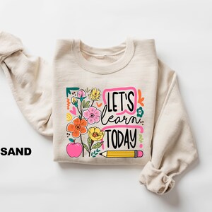 May include: Sand-colored sweatshirt with the text "LET'S learn TODAY" in a colorful design featuring flowers, an apple, and a pencil. The sweatshirt is folded with the sleeves rolled up.
