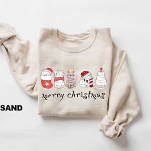 May include: A beige sweatshirt with a graphic of five cats wearing Christmas attire. The cats are lined up in a row and the text "merry christmas" is printed below them.