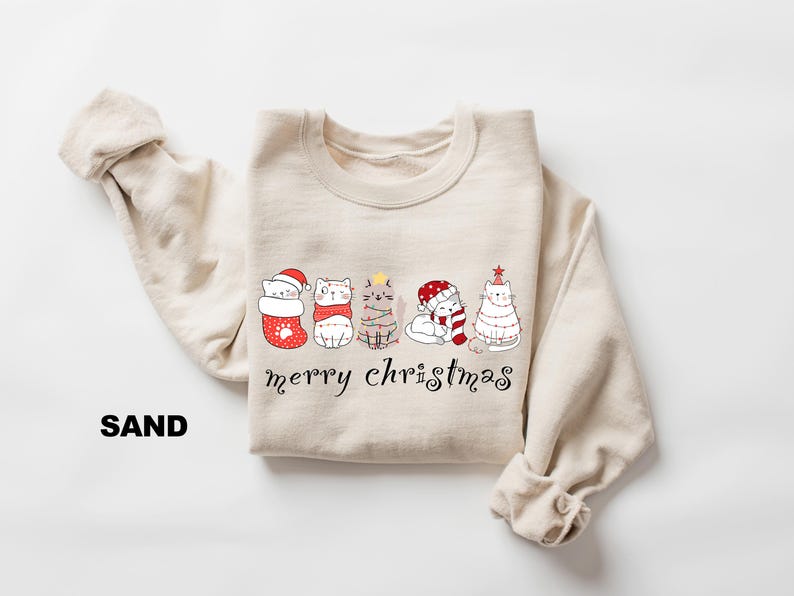 May include: A beige sweatshirt with a graphic of five cats wearing Christmas attire. The cats are lined up in a row and the text "merry christmas" is printed below them.