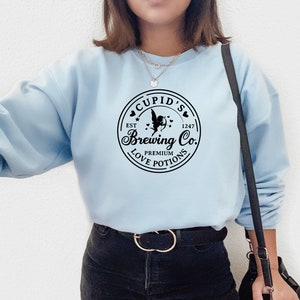 May include: A light blue sweatshirt with a black graphic of a logo that says "Cupid's Brewing Co. Est. 1247 Premium Love Potions" with a cupid holding an arrow and hearts around the logo.