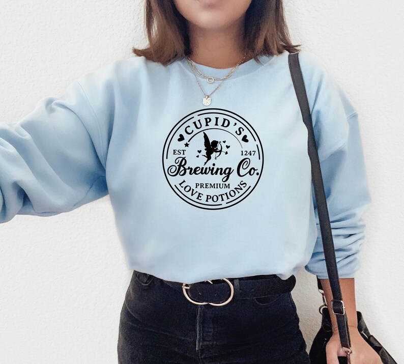 May include: A light blue sweatshirt with a black graphic of a logo that says "Cupid's Brewing Co. Est. 1247 Premium Love Potions" with a cupid holding an arrow and hearts around the logo.