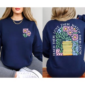 May include: Navy blue sweatshirt with a floral design. The front features a small embroidered bouquet of pink and yellow flowers. The back displays a larger floral arrangement with the text "Teach them, love them, watch them grow."