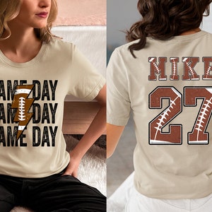 May include: Two beige t-shirts with a football theme. One shirt has the text "GAME DAY" repeated three times with a lightning bolt graphic made of a football. The other shirt has the text "MIKE" and the number "27" with a football graphic.