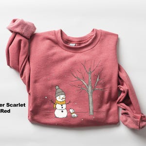May include: A heather scarlet red sweatshirt featuring a snowman and a dog illustration. The snowman is wearing a yellow scarf and a black hat. The dog is white with a black nose and tail.