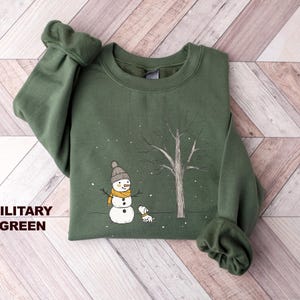 May include: A green sweatshirt featuring a snowman and a dog in a snowy forest scene. The text "MILITARY GREEN" is printed on the bottom left corner of the sweatshirt.
