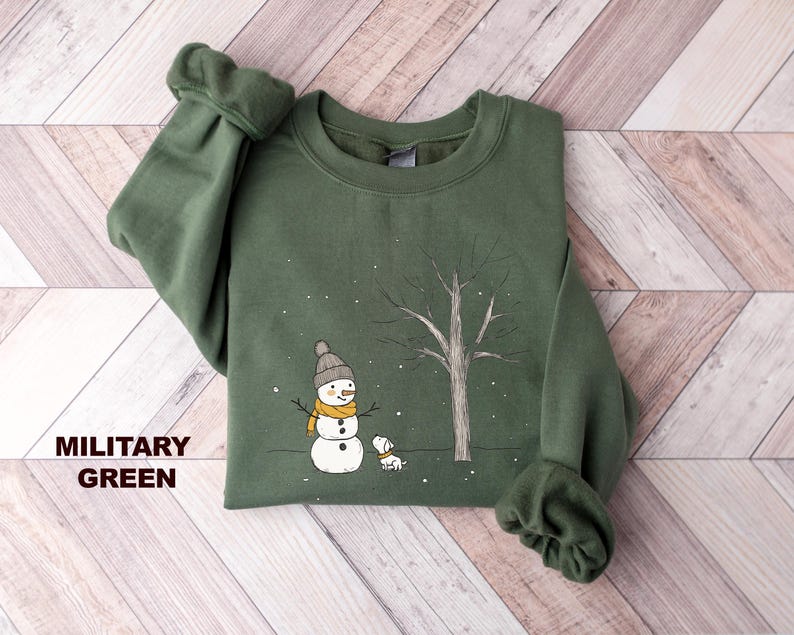 May include: A green sweatshirt featuring a snowman and a dog in a snowy forest scene. The text "MILITARY GREEN" is printed on the bottom left corner of the sweatshirt.