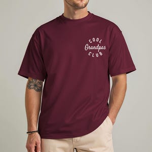 May include: A maroon t-shirt with the text "Cool Grandpas Club" in a white, curved font. The shirt has short sleeves and a crew neck. The model is wearing khaki pants and a bracelet.