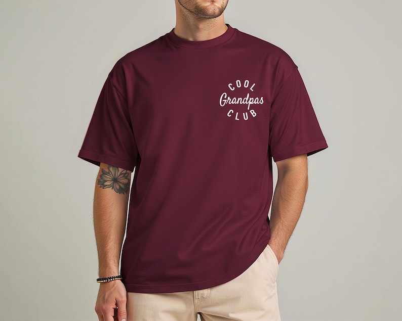 May include: A maroon t-shirt with the text "Cool Grandpas Club" in a white, curved font. The shirt has short sleeves and a crew neck. The model is wearing khaki pants and a bracelet.