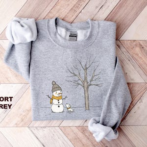May include: A grey sweatshirt featuring a snowman and a dog in a snowy scene. The snowman is wearing a yellow scarf and a black hat. The dog is white with a black nose. The text "SPORT GREY" is printed in the bottom left corner of the image.