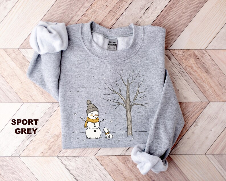 May include: A grey sweatshirt featuring a snowman and a dog in a snowy scene. The snowman is wearing a yellow scarf and a black hat. The dog is white with a black nose. The text "SPORT GREY" is printed in the bottom left corner of the image.