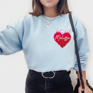 May include: Light blue sweatshirt with a red heart made of smaller hearts. The heart has the name "Melissa" written inside. The sweatshirt has a black strap and a black belt.