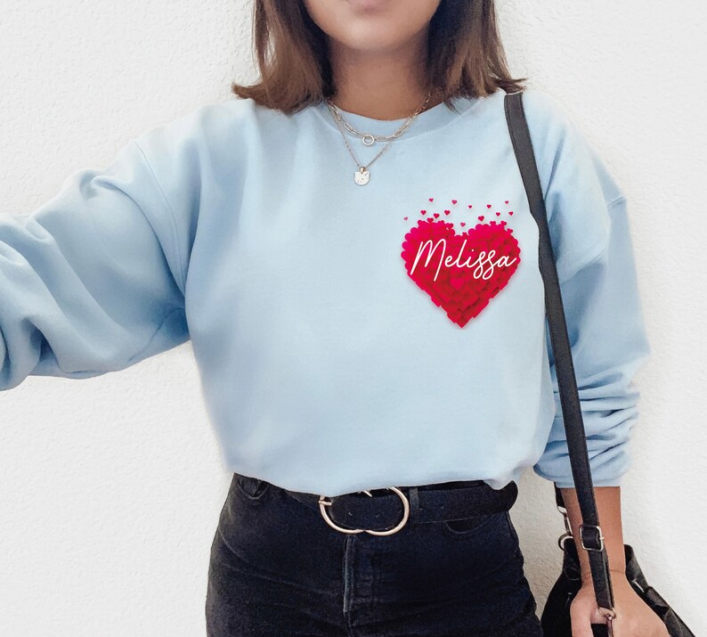 May include: Light blue sweatshirt with a red heart made of smaller hearts. The heart has the name "Melissa" written inside. The sweatshirt has a black strap and a black belt.