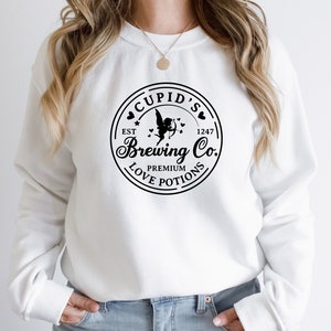 May include: White sweatshirt with a black circular logo that says "Cupid's Brewing Co." with a cupid design and the text "Est 1247 Premium Love Potions".