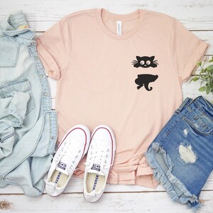 May include: A light pink t-shirt with a black silhouette of a cat peeking out from behind a pocket. The shirt is laid flat on a white wooden surface with a pair of white sneakers, a light blue denim jacket, and a pair of blue denim shorts.