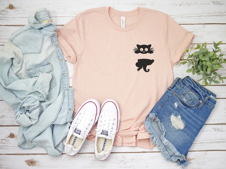 May include: A light pink t-shirt with a black silhouette of a cat peeking out from behind a pocket. The shirt is laid flat on a white wooden surface with a pair of white sneakers, a light blue denim jacket, and a pair of blue denim shorts.