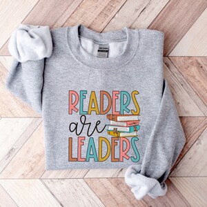 May include: Gray sweatshirt with the text "Readers are Leaders" in colorful letters. A stack of colorful books is behind the word "are".