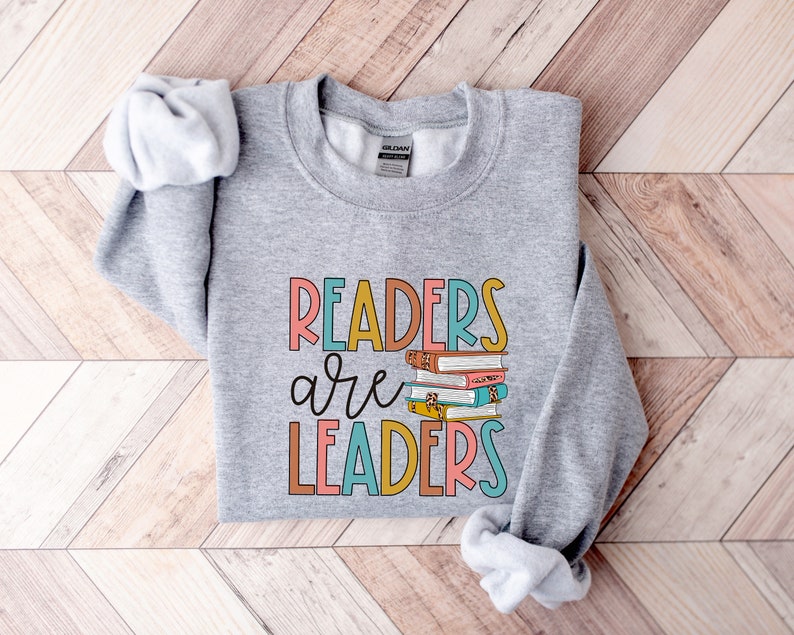 May include: Gray sweatshirt with the text "Readers are Leaders" in colorful letters. A stack of colorful books is behind the word "are".