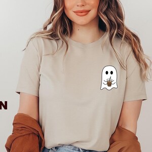 Puede incluir: Camiseta de color marr&oacute;n claro con un gr&aacute;fico de un fantasma blanco sosteniendo un caf&eacute; helado marr&oacute;n. El fantasma tiene dos ojos y una peque&ntilde;a sonrisa.