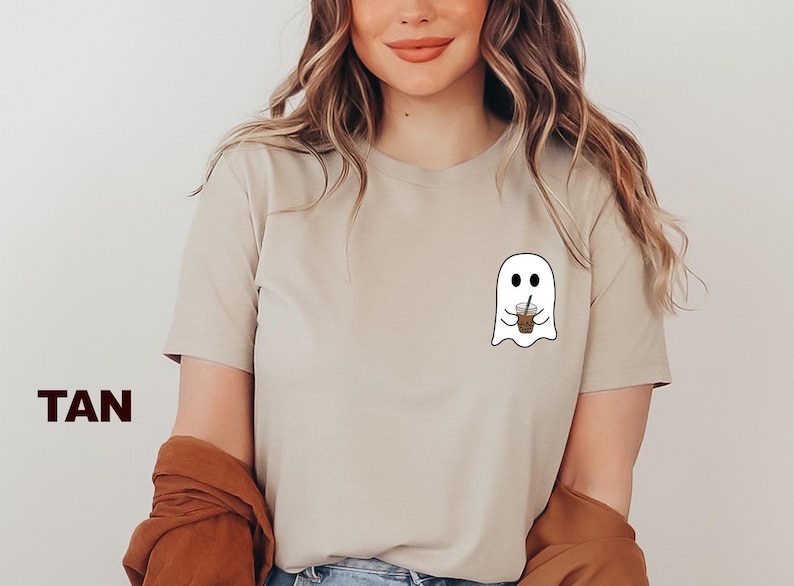 Puede incluir: Camiseta de color marr&oacute;n claro con un gr&aacute;fico de un fantasma blanco sosteniendo un caf&eacute; helado marr&oacute;n. El fantasma tiene dos ojos y una peque&ntilde;a sonrisa.