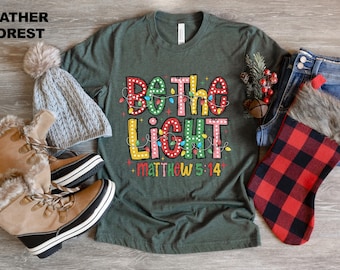 Op de afbeelding: Groen T-shirt met de tekst "Wees het licht" in kleurrijke letters met gloeilampen rond de woorden. De tekst "Mattheüs 5:14" staat onder de hoofdtekst.