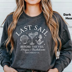 May include: Dark grey heather sweatshirt with a white graphic that reads "LAST SAIL BEFORE THE VEIL" with seashell, cocktail, and anchor illustrations. Below the text is "Megan's Bachelorette, San Diego, CA".