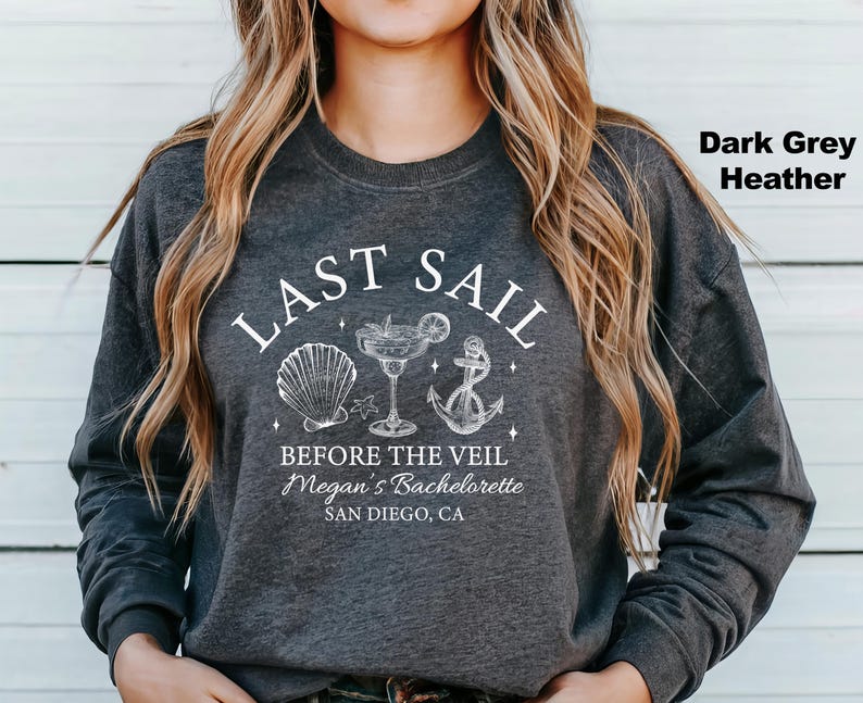 May include: Dark grey heather sweatshirt with a white graphic that reads "LAST SAIL BEFORE THE VEIL" with seashell, cocktail, and anchor illustrations. Below the text is "Megan's Bachelorette, San Diego, CA".