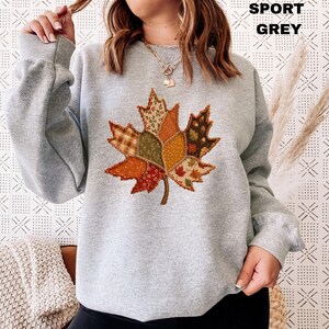 Pode incluir: Uma camisola de manga comprida em cinzento desportivo com um grande gr&aacute;fico central de uma folha de &aacute;cer composta por v&aacute;rios padr&otilde;es de tecido patchwork em tons de laranja, castanho, verde e amarelo. O texto SPORT GREY est&aacute; impresso no canto superior direito.