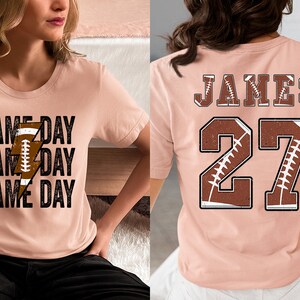 May include: Two pink t-shirts with a football theme. One shirt has the text "GAME DAY" repeated three times with a football and lightning bolt graphic. The other shirt has the name "JAMES" and the number "27" with a football graphic.