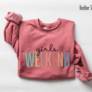 Peut inclure: Un sweat-shirt rouge &eacute;carlate chin&eacute; avec le texte "girls weekend" en couleurs arc-en-ciel.