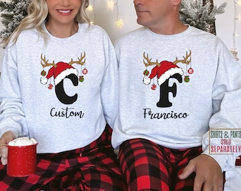 Pode incluir: Um par de suéteres de Natal cinzentos combinando com um design de chifres de rena e um chapéu de Papai Noel com uma letra e um nome. Os suéteres são usados por um casal. O suéter da mulher tem a letra "C" e o nome "Custom". O suéter do homem tem a letra "F" e o nome "Francisco".