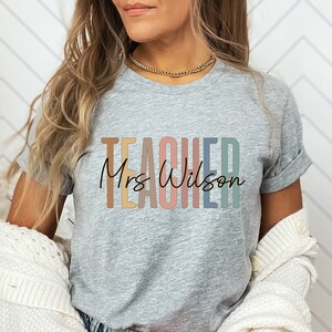 K&ouml;nnte beinhalten: Ein graues T-Shirt mit dem Wort "TEACHER" in Regenbogenfarben und dem Text "Mrs. Wilson" in schwarzer Schrift.