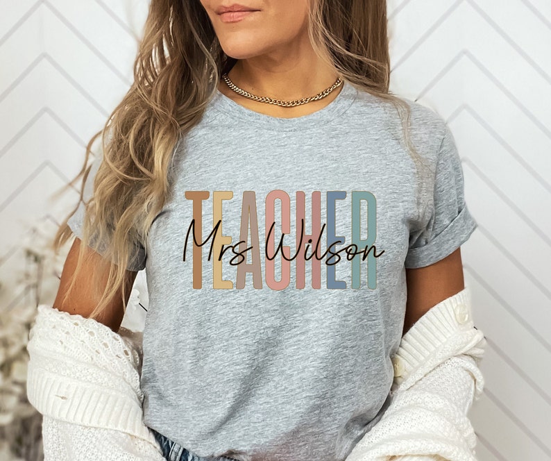 K&ouml;nnte beinhalten: Ein graues T-Shirt mit dem Wort "TEACHER" in Regenbogenfarben und dem Text "Mrs. Wilson" in schwarzer Schrift.
