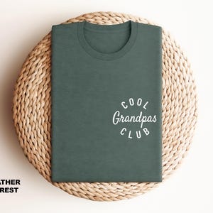 May include: A heather forest green t-shirt with the text "Cool Grandpas Club" in white script. The shirt is folded and placed on a woven, circular surface. The words "Heather Forest" are printed at the bottom.