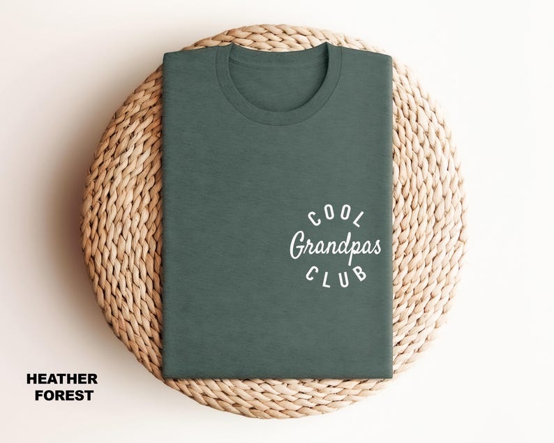 May include: A heather forest green t-shirt with the text "Cool Grandpas Club" in white script. The shirt is folded and placed on a woven, circular surface. The words "Heather Forest" are printed at the bottom.